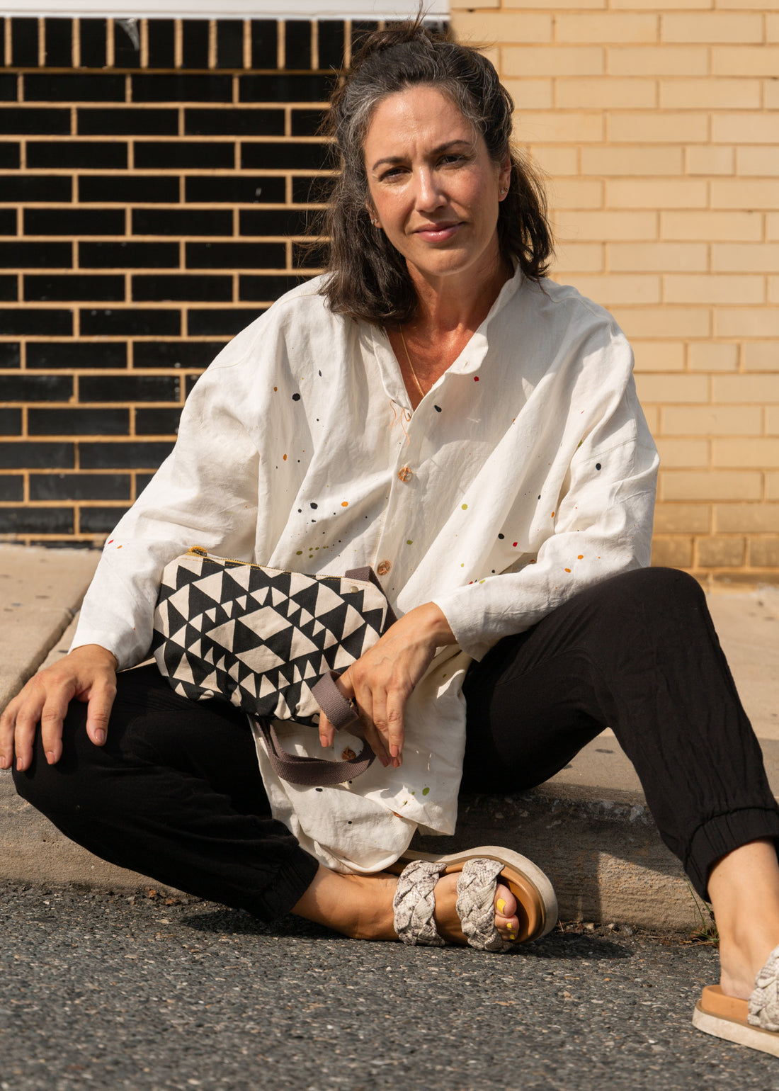 a woman sitting on sidewalk looking at camera with an aztec styled bag in cream and black in her lap and a strap wrapped around her finger