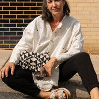 a woman sitting on sidewalk looking at camera with an aztec styled bag in cream and black in her lap and a strap wrapped around her finger