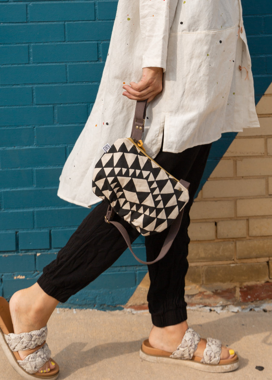 a woman sitting walking down a sidewalk with an aztec styled bag in cream and black in her hand and a strap wrapped around her finger