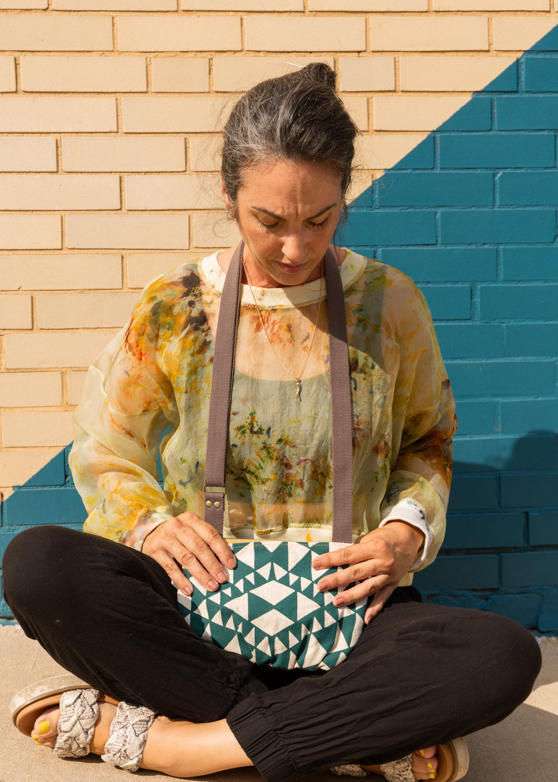 a woman sitting in front of a brick wall with an aztec designed crossbody bag around her neck