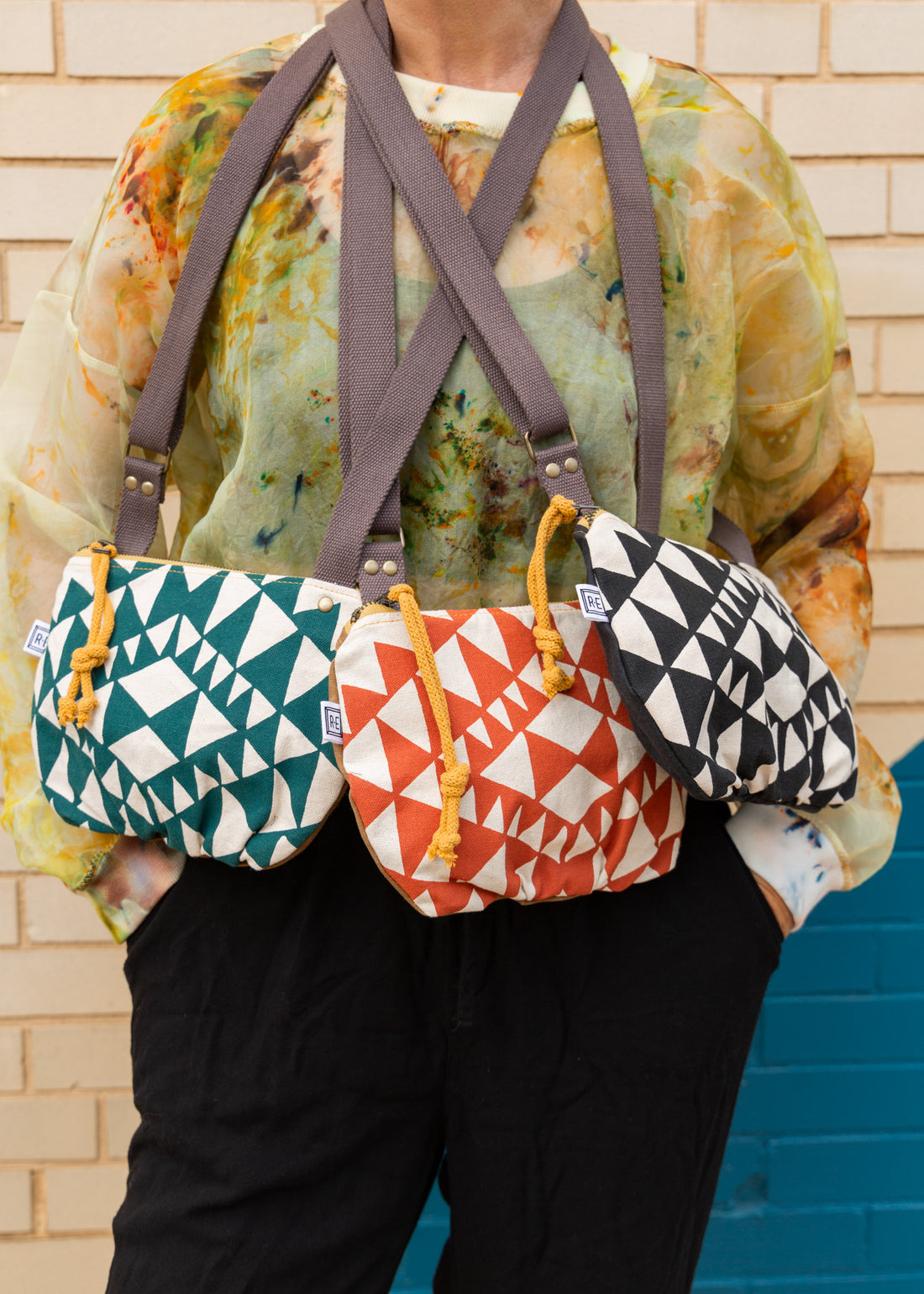 a woman in front of a brick wall with three crossbody bags hanging from her neck, all in different color variations