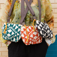 a woman in front of a brick wall with three crossbody bags hanging from her neck, all in different color variations