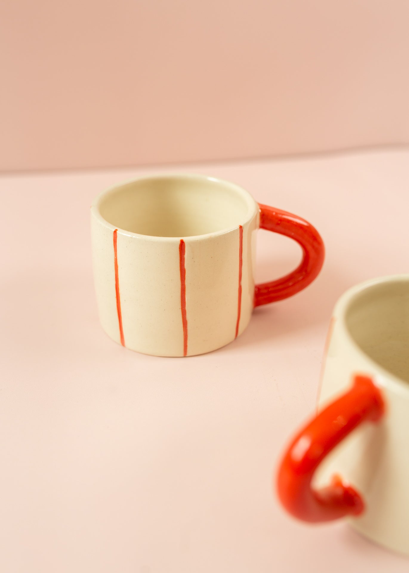 Photo of a cream ceramic mug with light red stripes going around the mug vertically, and a red handle. Another mug is in the foreground