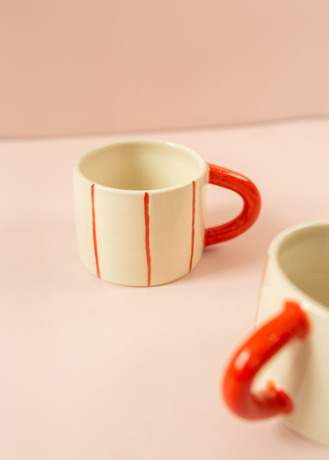 Photo of a cream ceramic mug with light red stripes going around the mug vertically, and a red handle. Another mug is in the foreground