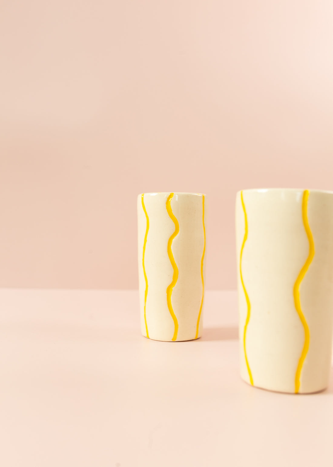 Smal cream vase with squiggly, bright yellow paint lines running vertically around the vase. Another vase in the foreground