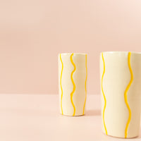Smal cream vase with squiggly, bright yellow paint lines running vertically around the vase. Another vase in the foreground