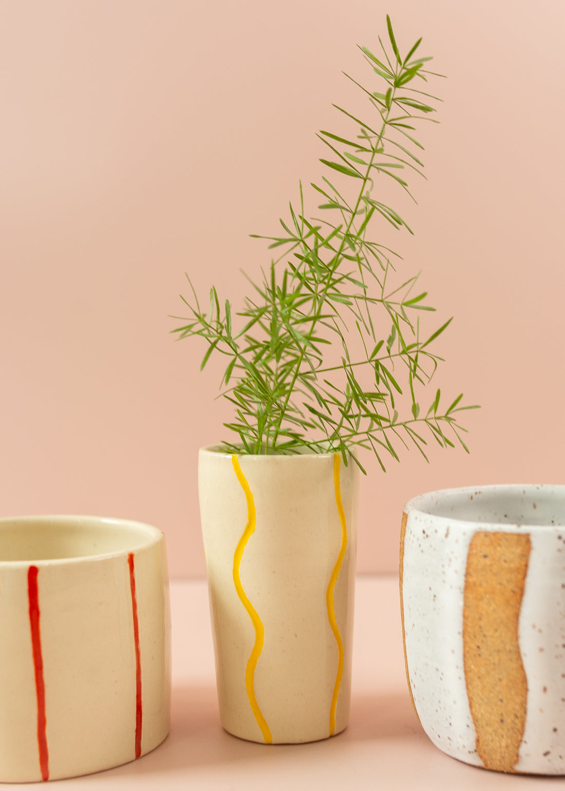 Smal cream vase with squiggly, bright yellow paint lines running vertically around the vase. There is a green branch in the vase, and two mugs on either side of vase.