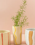 Smal cream vase with squiggly, bright yellow paint lines running vertically around the vase. There is a green branch in the vase, and two mugs on either side of vase.