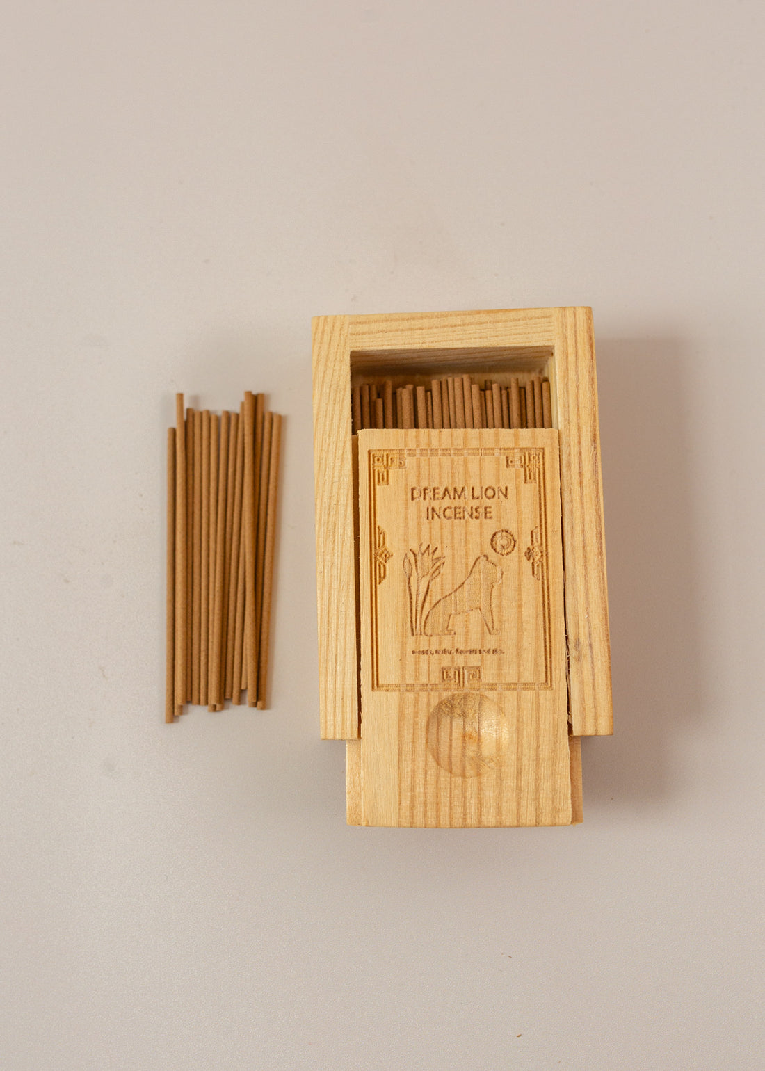 A wooden box laying on pink background, half open to reveal mini incense sticks with a few piked up to the left of the box.