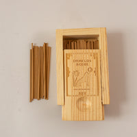 A wooden box laying on pink background, half open to reveal mini incense sticks with a few piked up to the left of the box.