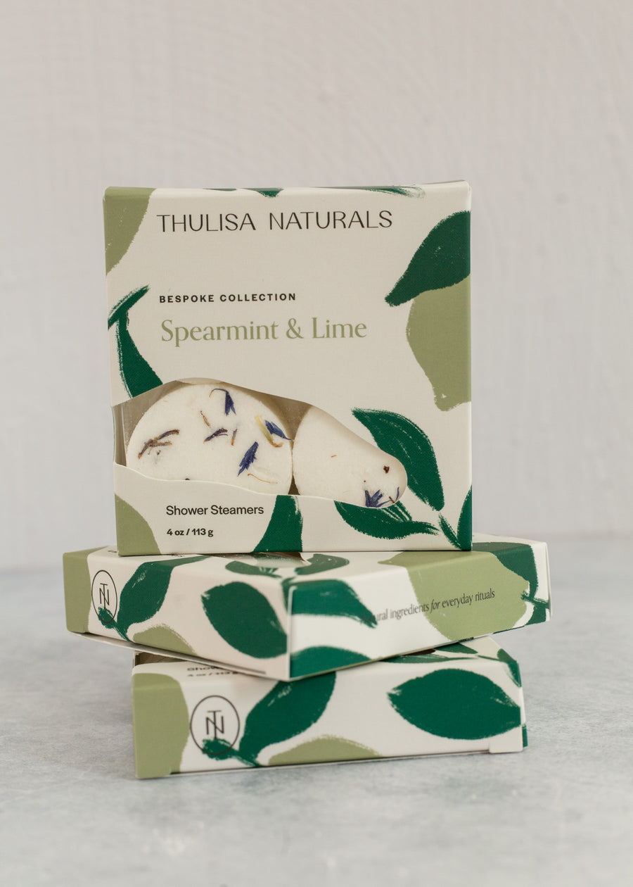Two boxes of shower steamers with one standing up on top. Boxes are white and green and "spearmint and lime" scented