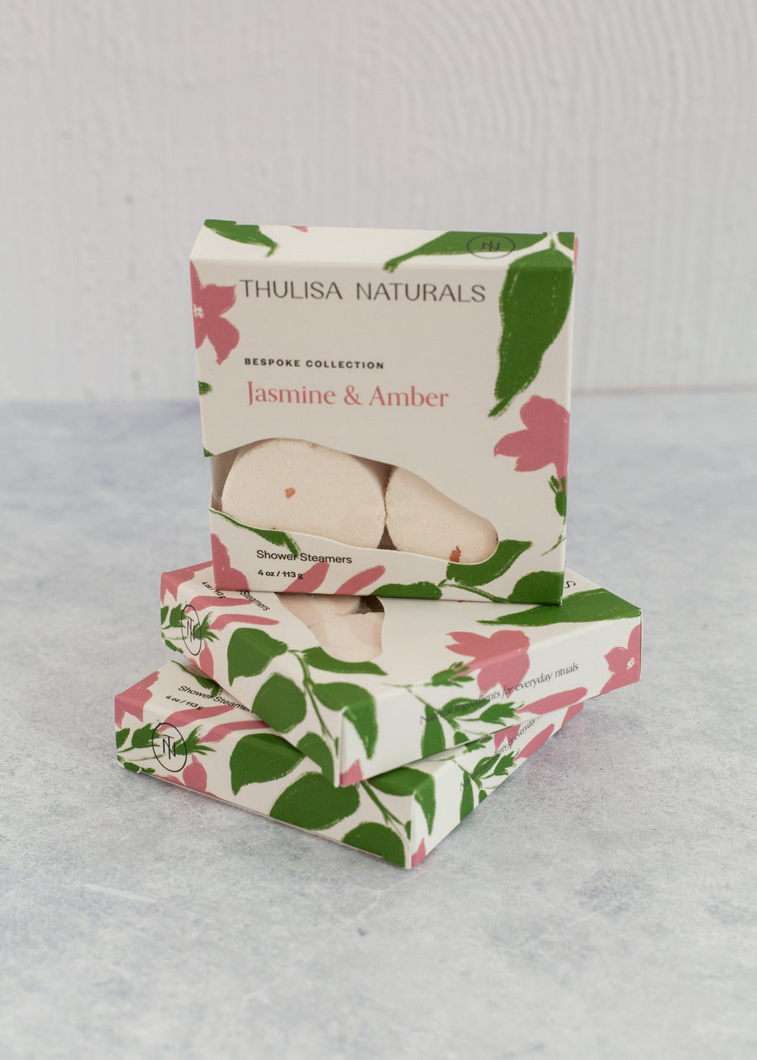 Two boxes of shower steamers with one standing up on top. Boxes are white, pink and green and "jasmine and amber" scented