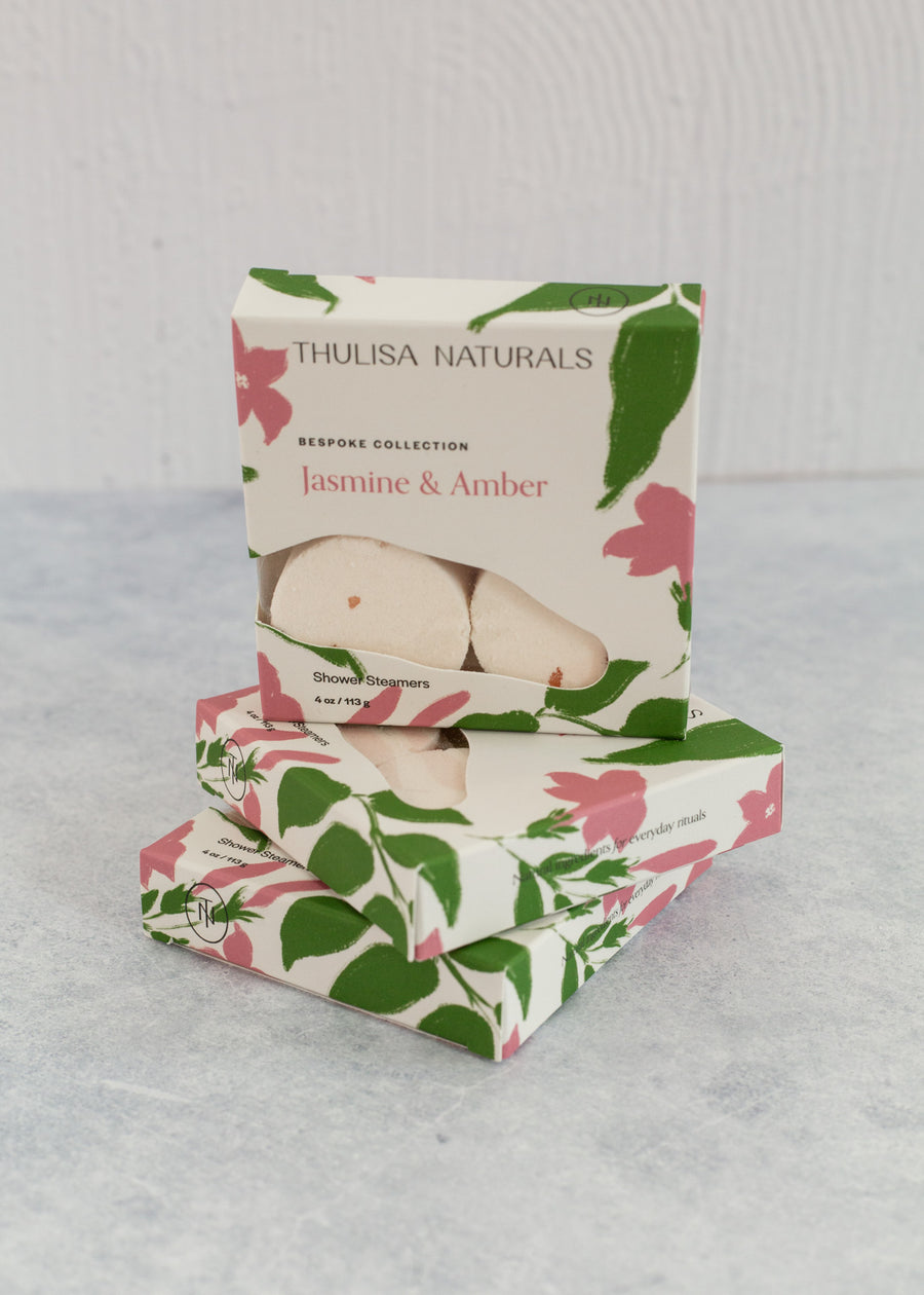 Two boxes of shower steamers with one standing up on top. Boxes are white, pink and green and "jasmine and amber" scented