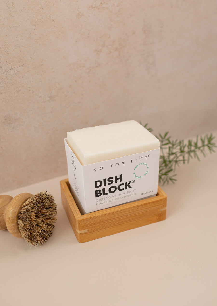 Dish soap block sitting in a bamboo soap shelf, all on a light pink background with a dish brush and green branch in the background