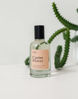 Perfume bottle on a white background with a cactus in the background and with a light pink label saying "Cactus Flower"