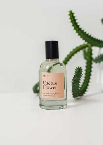 Perfume bottle on a white background with a cactus in the background and with a light pink label saying "Cactus Flower"