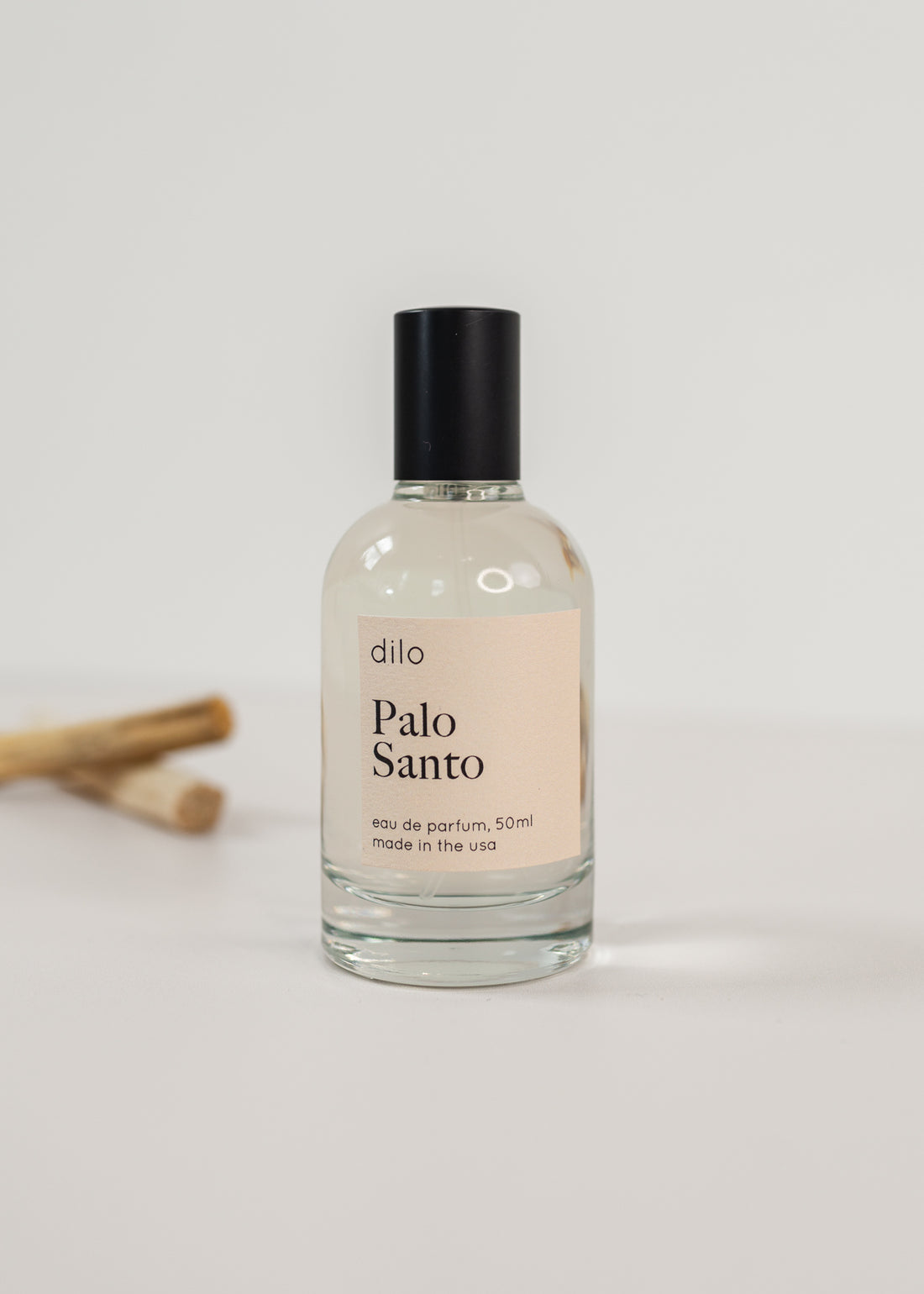 Perfume bottle on a white background with two sticks on palo santo in the background with a light pink label saying "Palo Santo"