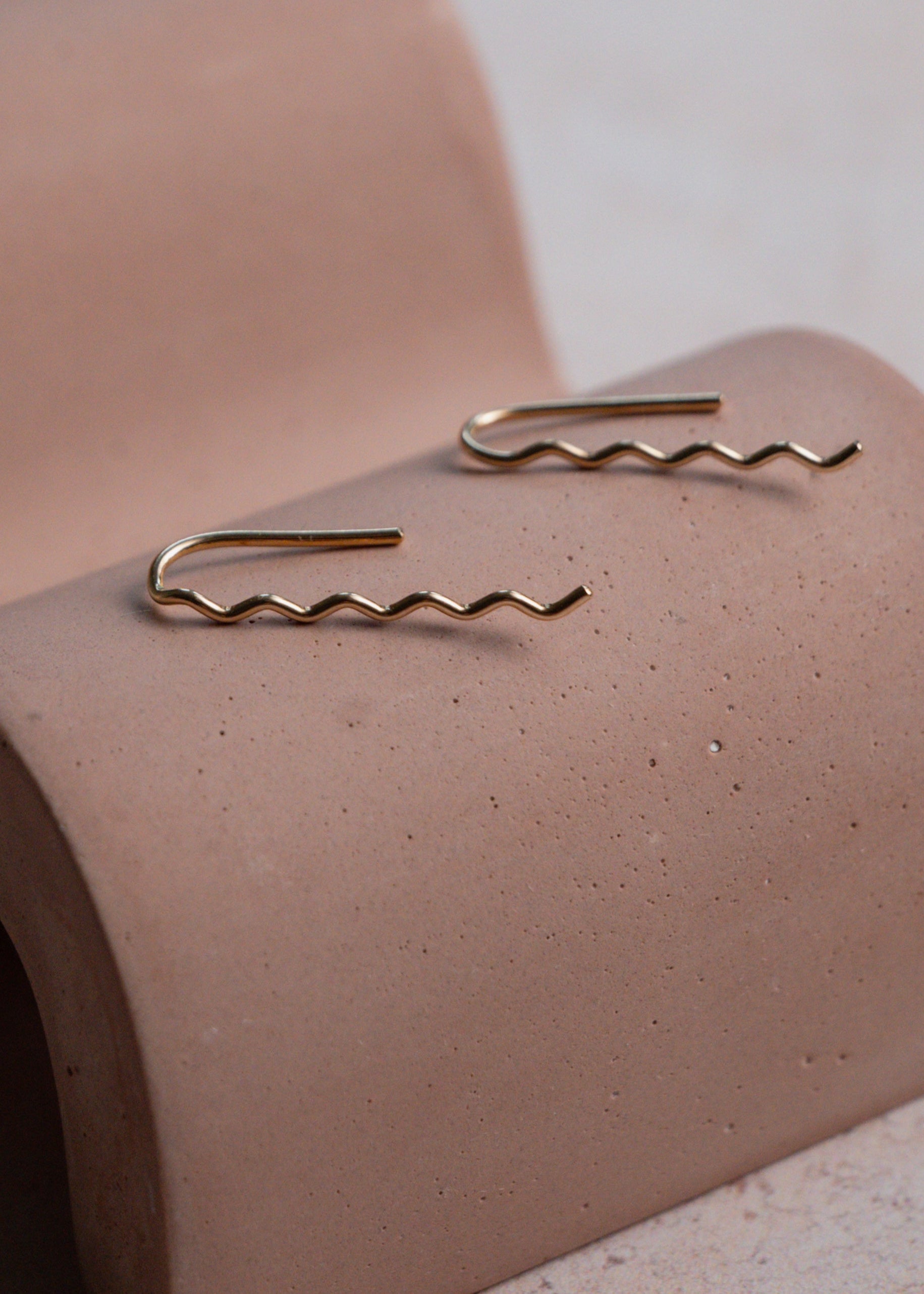 Two gold wavy earrings on a wavy display object with a neutral background