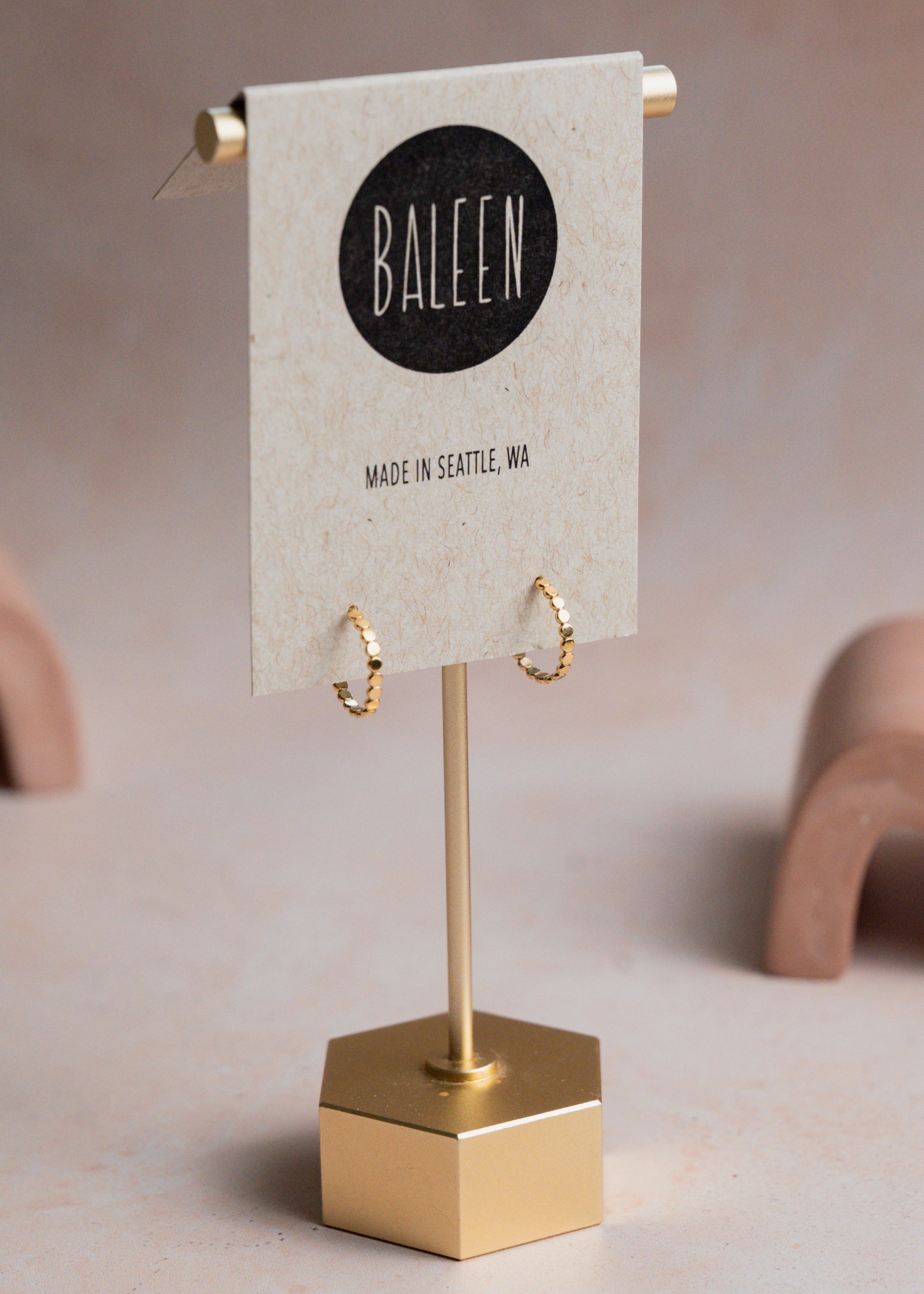 Gold earrings on a stand with 'Baleen' brand card on a beige background