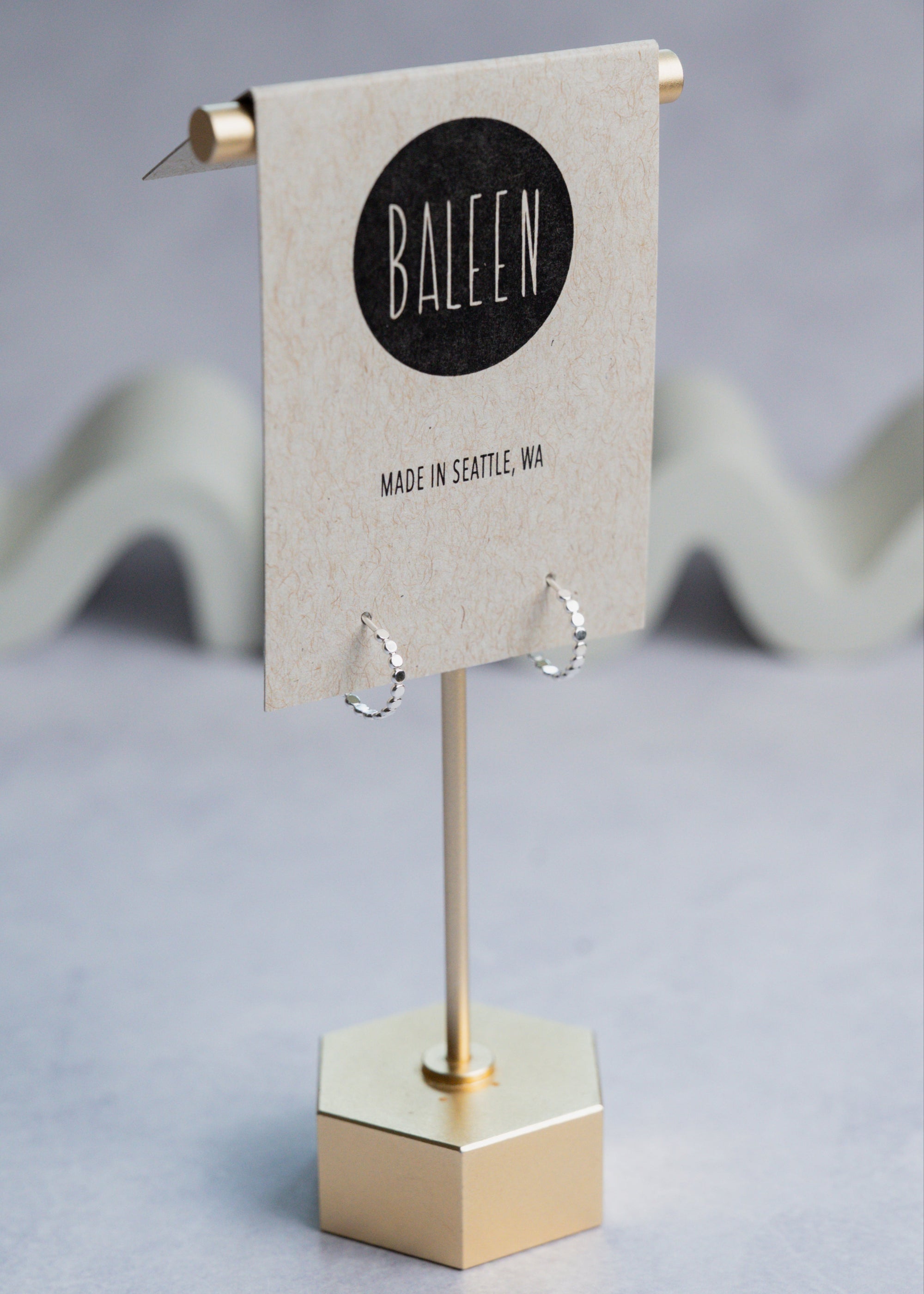 Silver hoop earrings on a stand with 'Baleen' brand label on a gray background