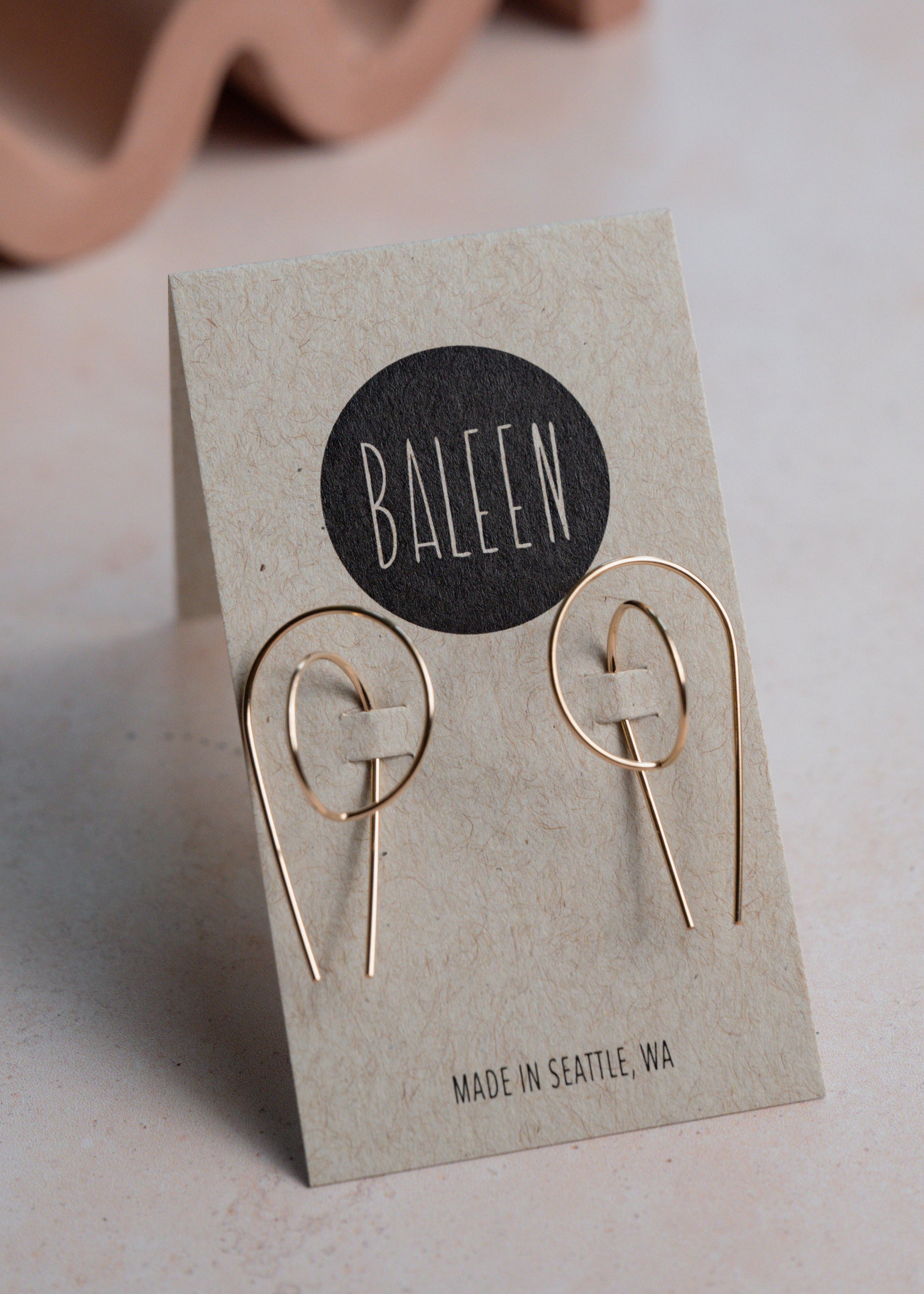 Gold earrings on a 'BALEEN' branded card with a wavy object in the background
