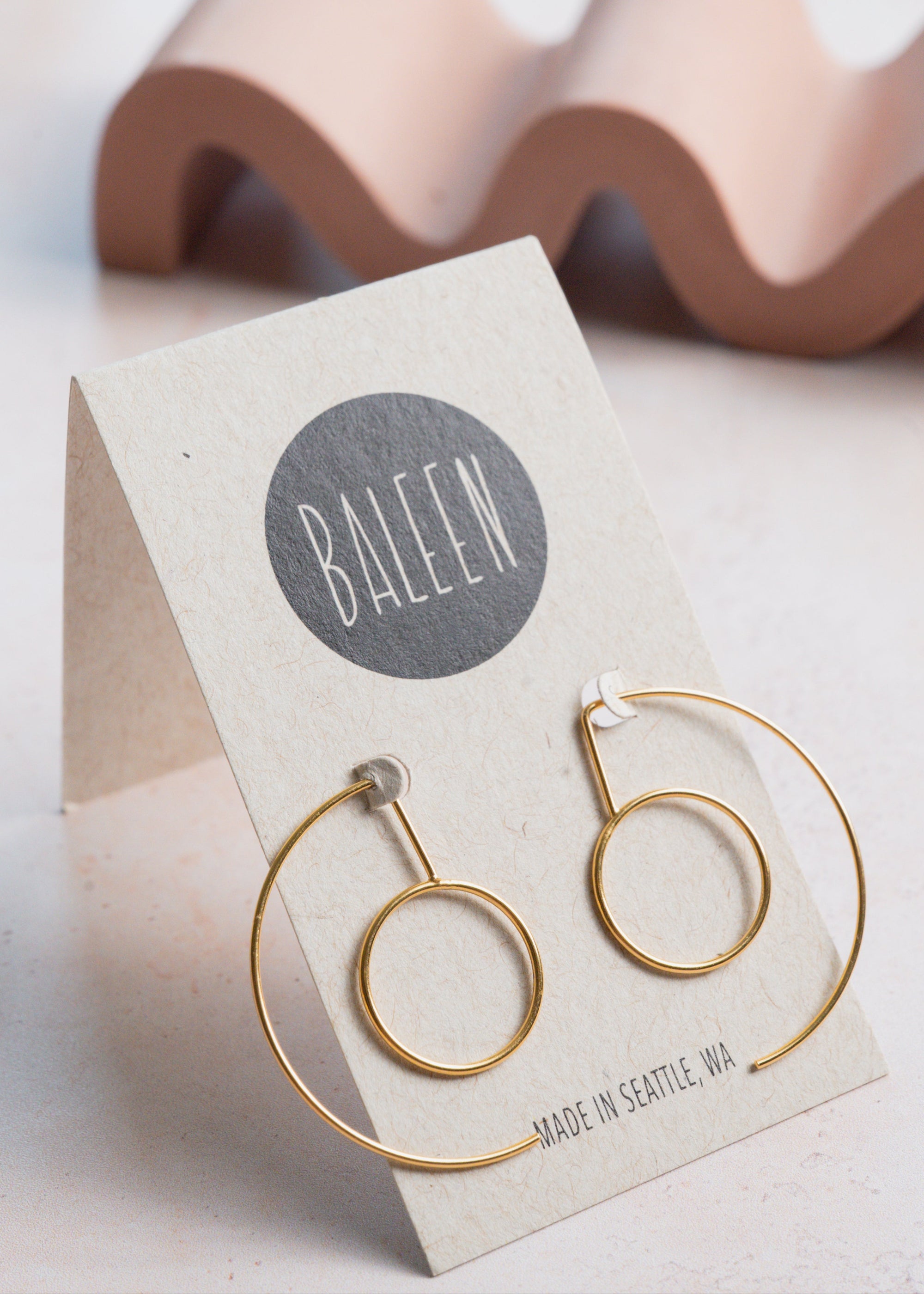 Gold hoop earrings on a BALEEN branded card with a wavy object in the background.