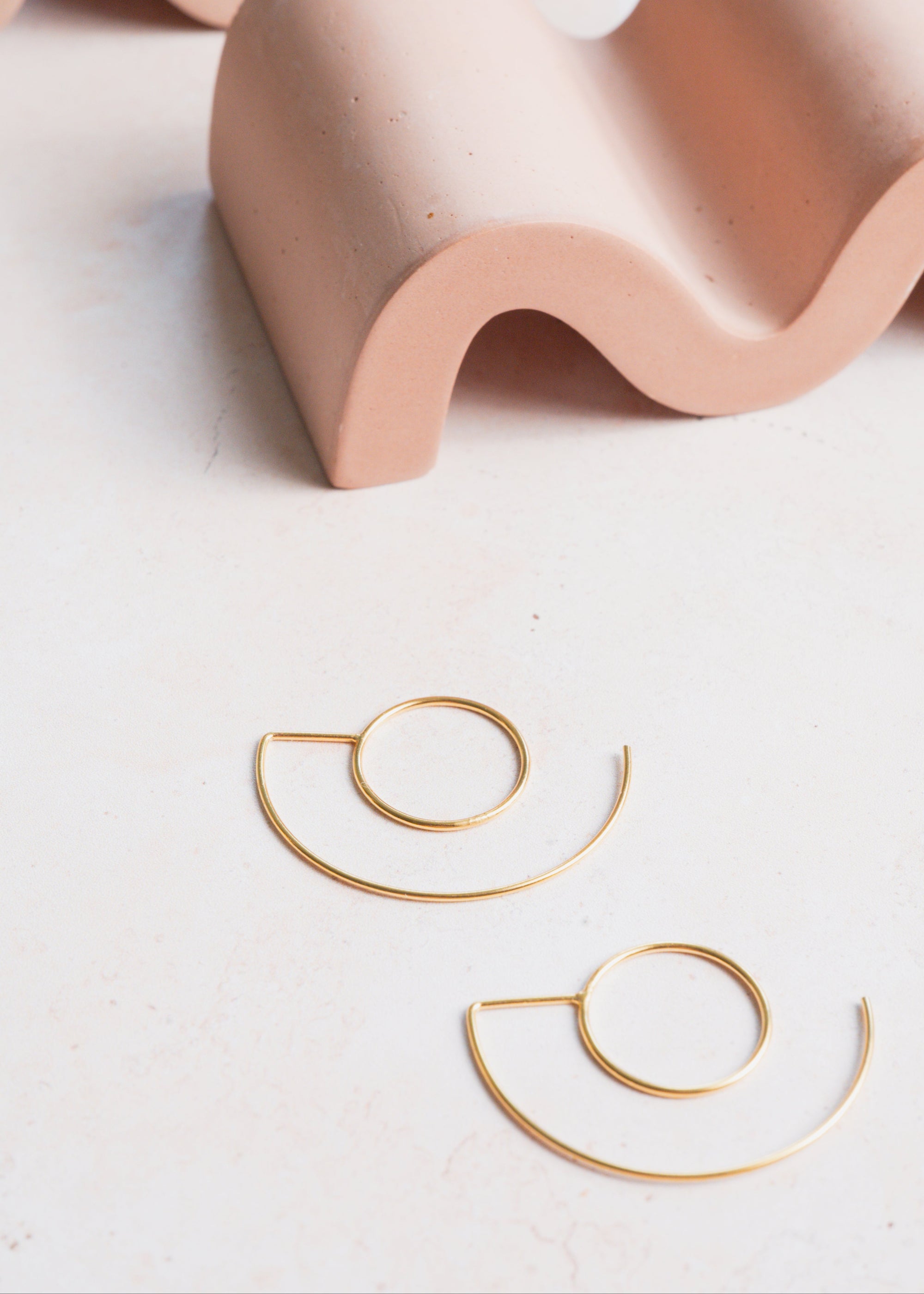 Gold hoop earrings on a light surface with a wavy pink ceramic holder in the background.