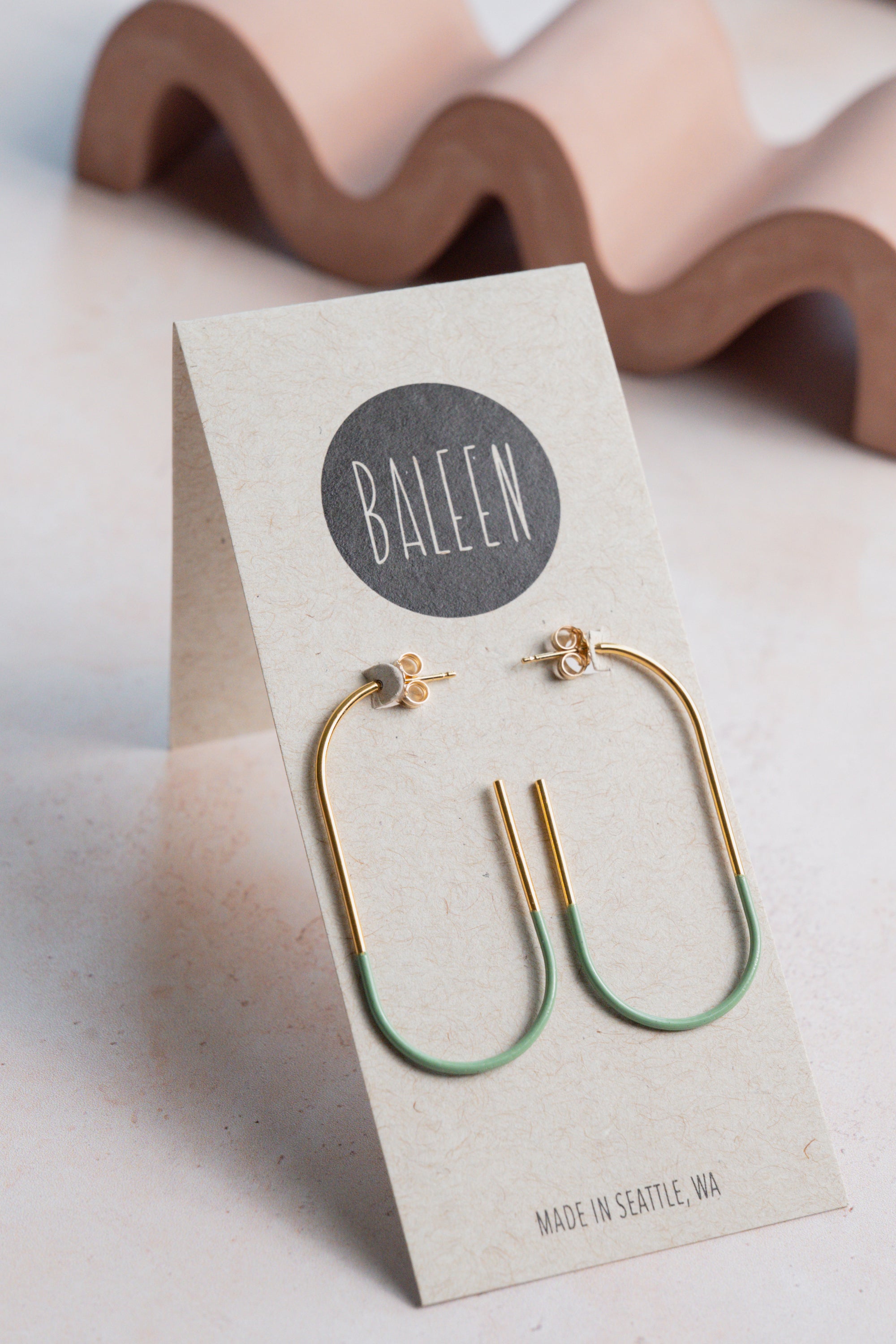 Gold and green earrings on a 'BALEEN' branded card with a wavy object in the background.