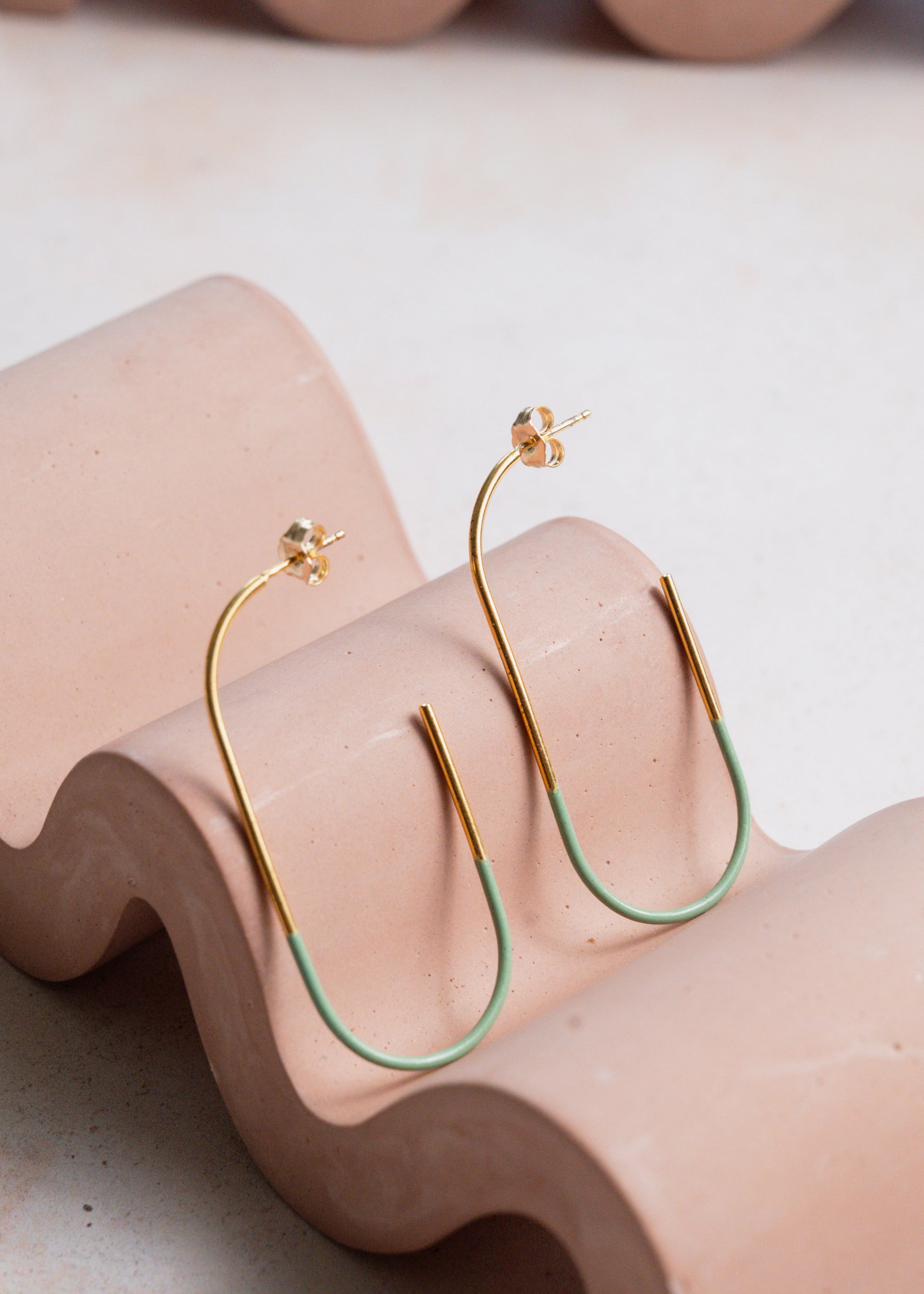 Gold and mint hoop earrings on a wavy pink stand with a neutral background