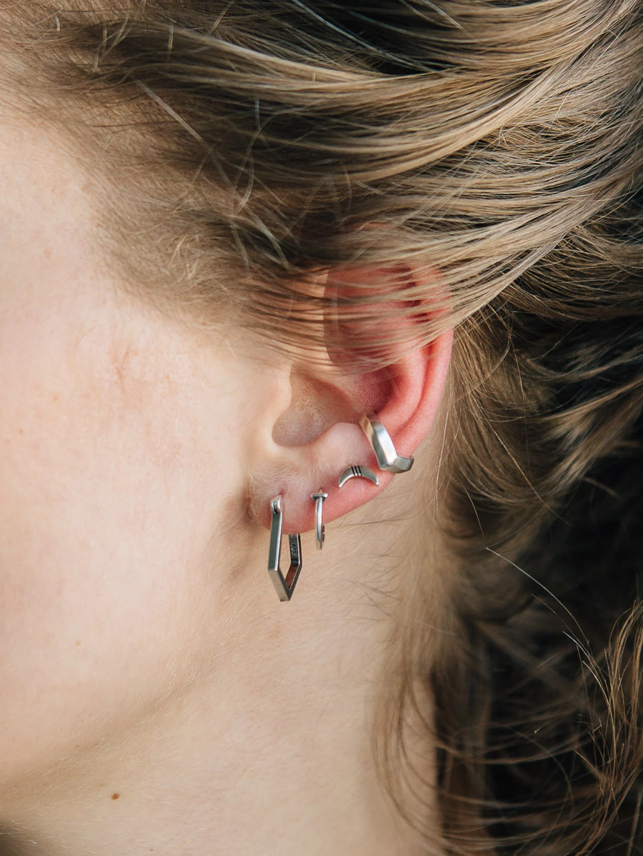 close-up of models ear with four types of sterling silver earrings. Front left to right: Pentagon shaped earring, simple hoop, big sky crescent moon stud, and a silver cuff.