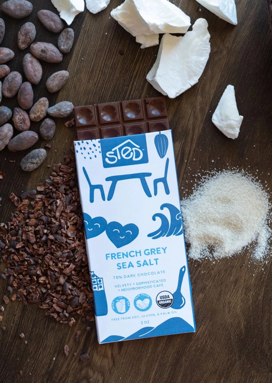 on a dark wooden table, a french grey sea salt bar with square pieces coming out of the top with cacao beans, crushed chocolate, coconut and sea salt scattered around it.