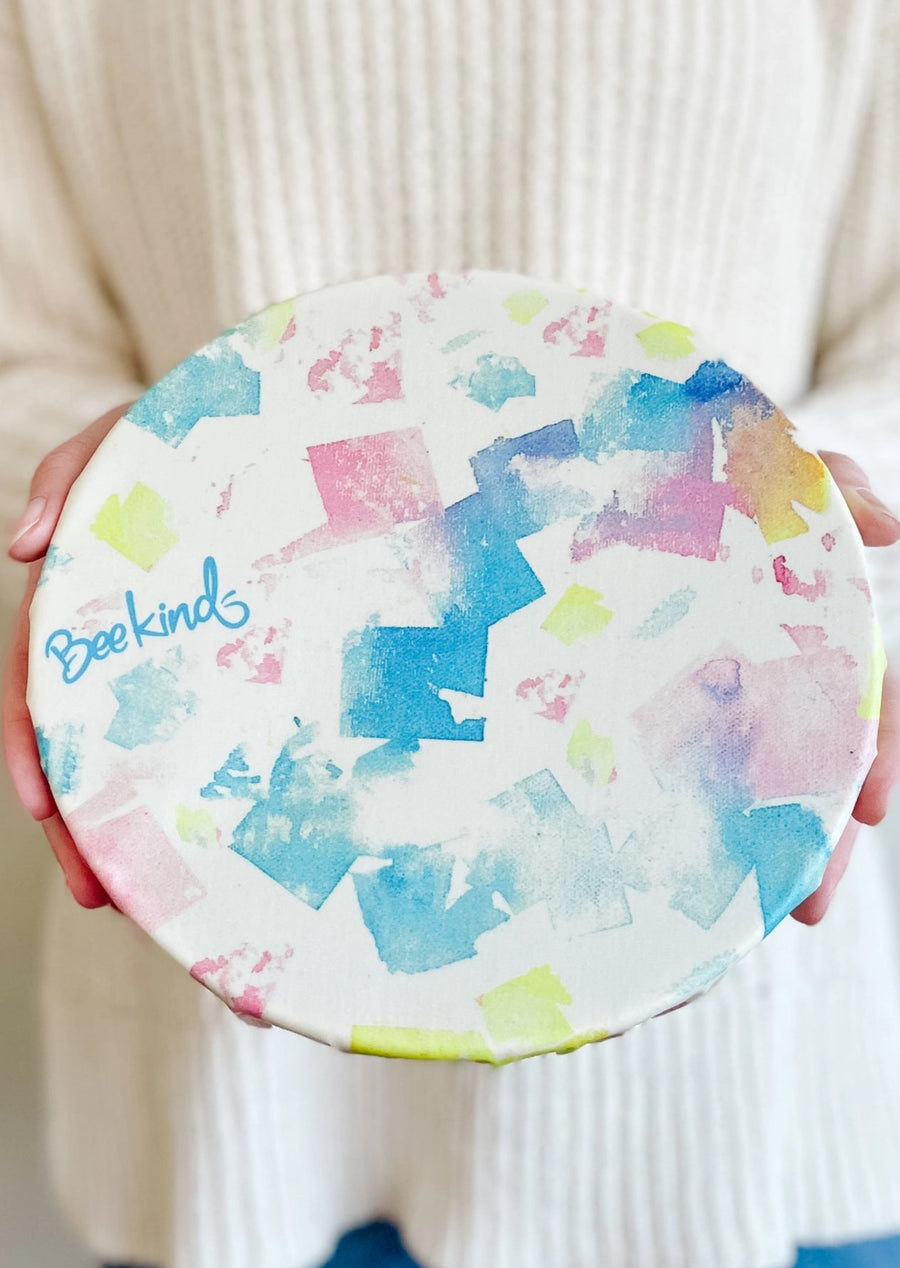 off-white colored beeswax wrap with watercolored-like square patterns in variety of colors like blue, light green and pink, covering a bowl with a lady holding the bowl.