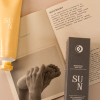 Flatlay of a mini yellow tube on botanical body cream named 'sun' next to the box it comes in, with a paper underneath with guides and techniques on how to apply.