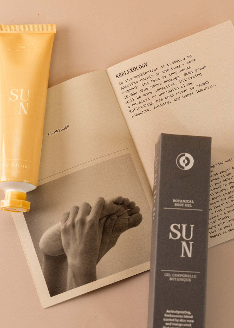 Flatlay of a mini yellow tube on botanical body cream named 'sun' next to the box it comes in, with a paper underneath with guides and techniques on how to apply.