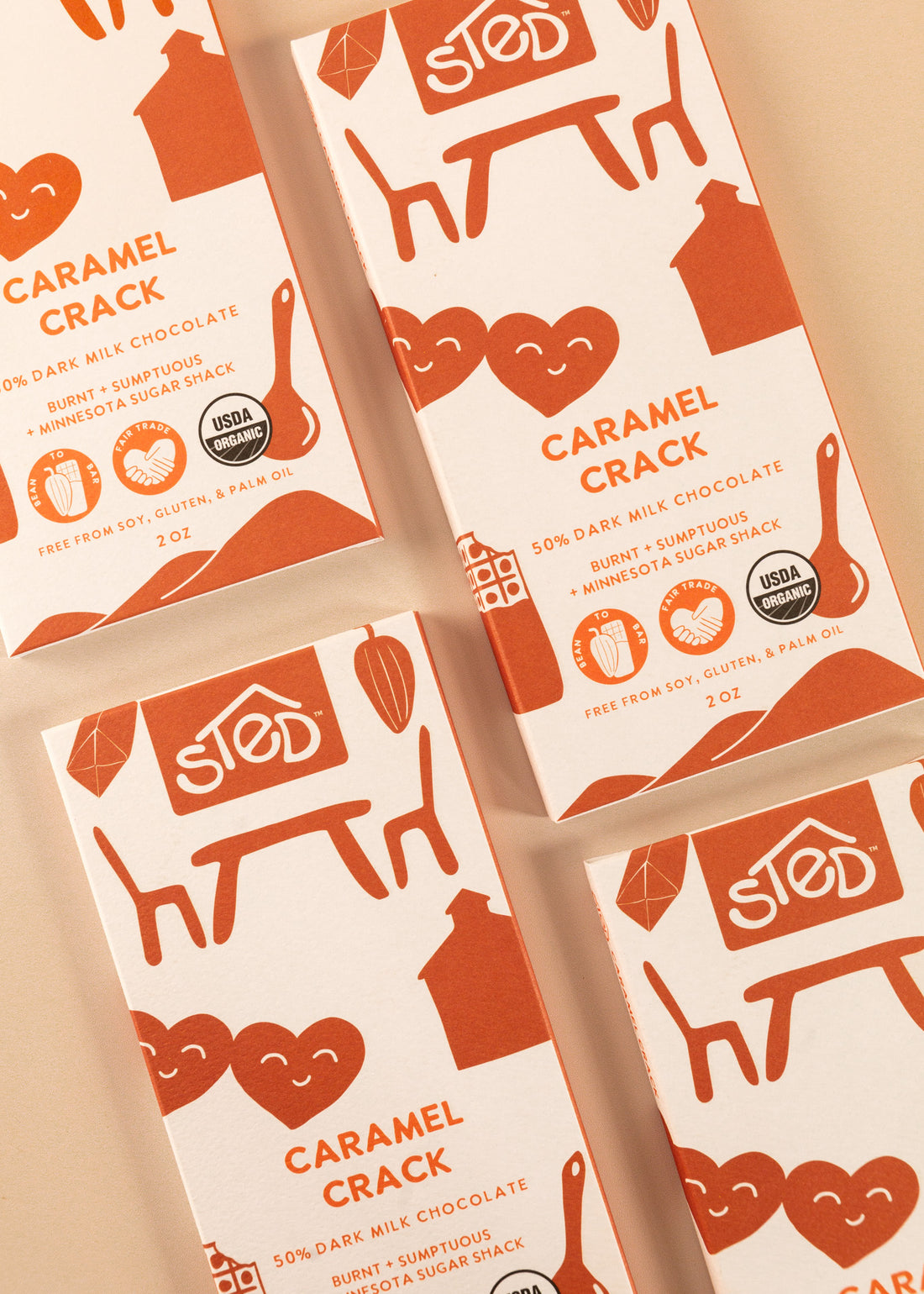 flatlay of four covers of a 50% dark milk chocolate bar by Sted. A white cover with deep orange artwork throughout, named ‘caramel crack’