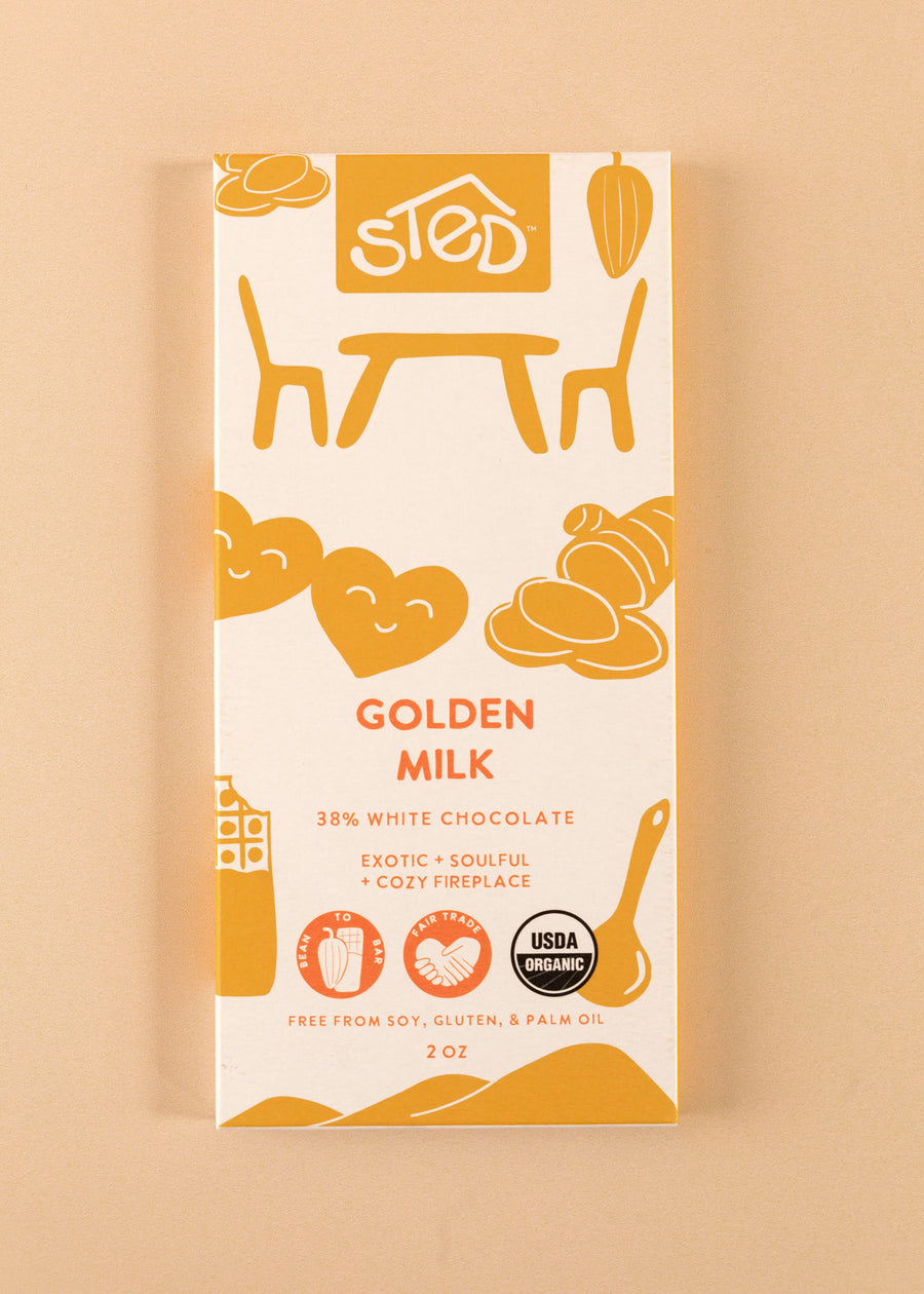 flatlay of the cover of a 38% white chocolate bar by Sted. A white cover with light orange artwork throughout, named 'golden milk'