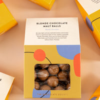 Flatly of house-shaped orange boxes with fun shapes throughout. Front of the box is a small window to see the malt balls, and the top states “blonde chocolate malt balls. Blonde chocolate. We take light and airy malt ball centers and enrobe them in a generous layer of rich, creamy, caramel blonde chocolate. Once you try these addictive little treats, you’ll wonder how you ever lived without them. Promise.”