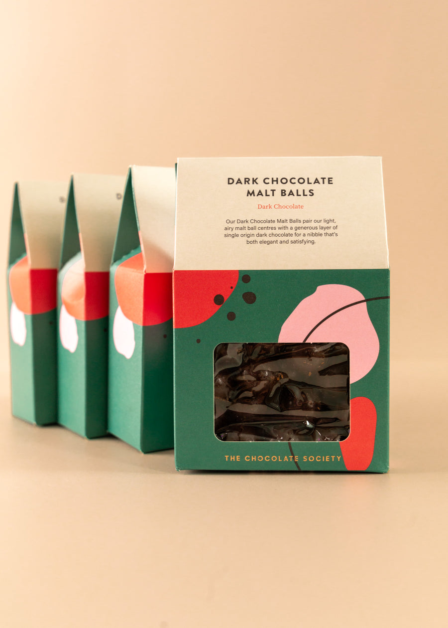 Four boxes of house-shaped green boxes with fun shapes throughout. Front of the box is a small window to see the malt balls, and the top states “dark chocolate malt balls. Dark chocolate. Our dark chocolate malt balls pair our light, airy malt ball centers with a generous layer of single origin dark chocolate for a nibble that’s both elegant and satisfying”