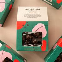 Flatly of house-shaped green boxes with fun shapes throughout. Front of the box is a small window to see the malt balls, and the top states “dark chocolate malt balls. Dark chocolate. Our dark chocolate malt balls pair our light, airy malt ball centers with a generous layer of single origin dark chocolate for a nibble that’s both elegant and satisfying”