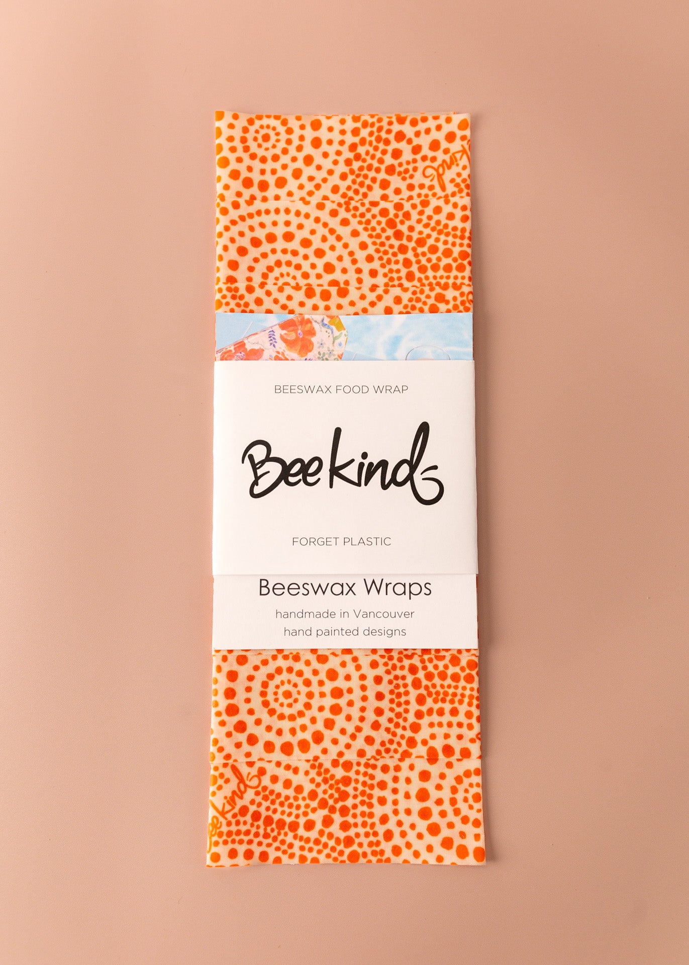 Set of 3 beeswax wraps that are light orange with darker prange dots throughout in a circle pattern, folded with a white label saying &quot;beekind&quot; on a pink flatlay