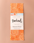 Set of 3 beeswax wraps that are light orange with darker prange dots throughout in a circle pattern, folded with a white label saying "beekind" on a pink flatlay