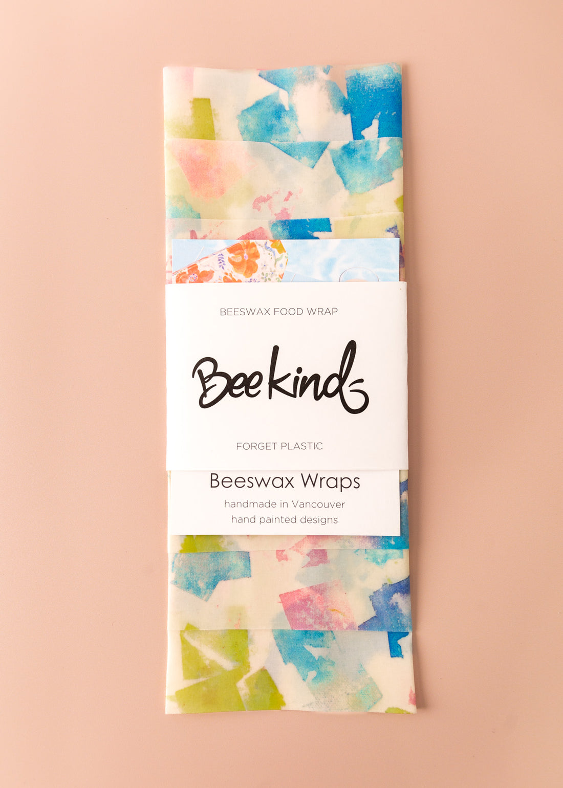 Set of 3 beeswax wraps that are off-white colored with watercolored-like square patterns in variety of colors like blue, light green and pink, folded with a white label saying "beekind" on a pink flatlay