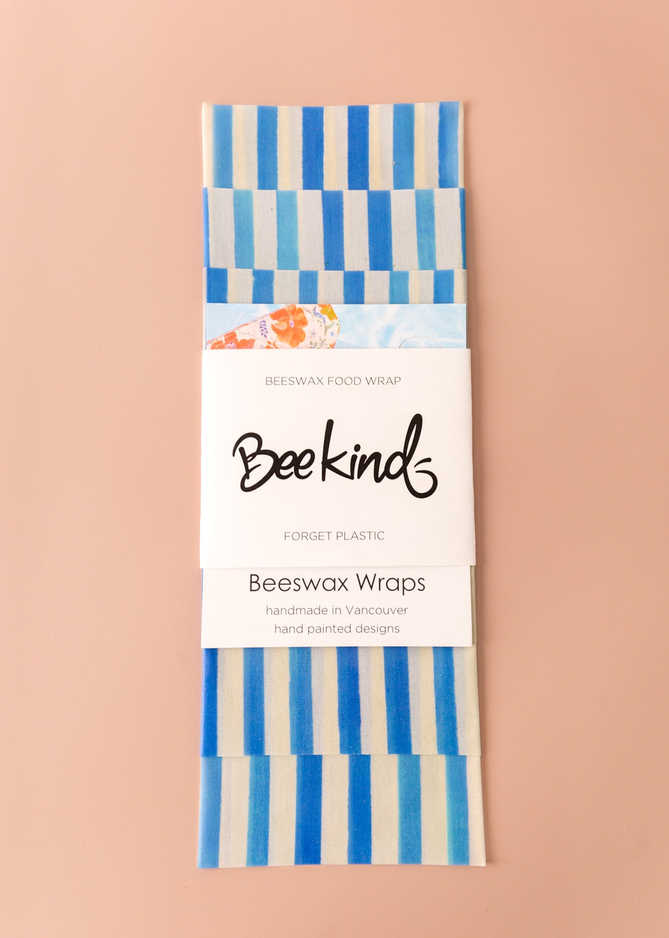 Set of 3 beeswax wraps that are off-white with thick blue stripes throughout, folded with a white label saying &quot;beekind&quot; on a pink flatlay