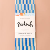 Set of 3 beeswax wraps that are off-white with thick blue stripes throughout, folded with a white label saying "beekind" on a pink flatlay