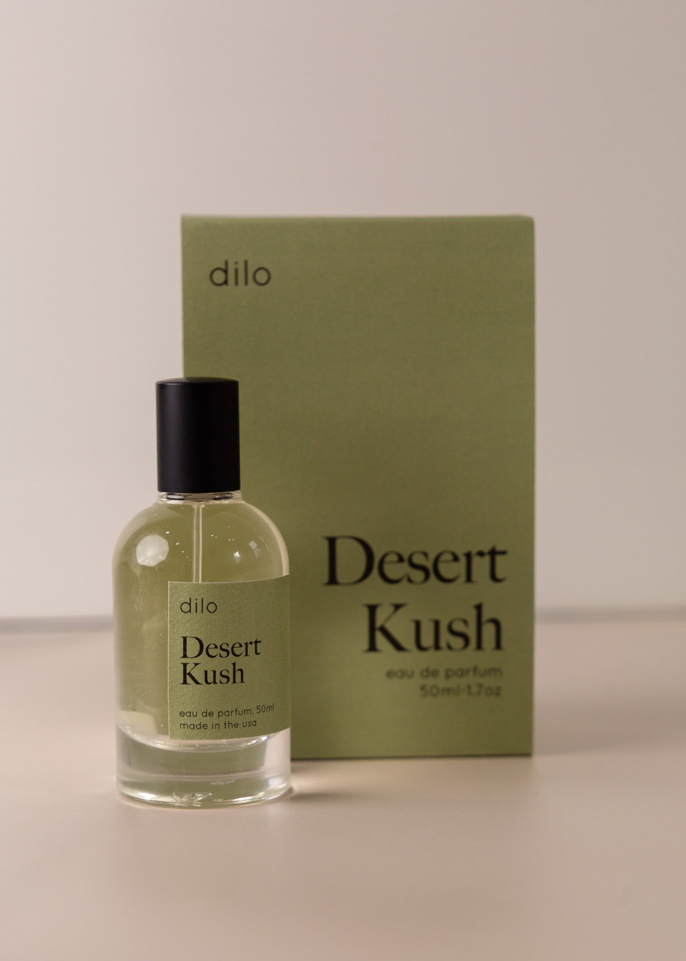 Bottle of perfume by dilo, with a green label on the front stating “desert kush. eau de parfum, 50 ml. made in the usa" with the green box that it comes in in the background
