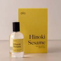 Bottle of perfume by dilo, with a yellow label on the front stating "hinoki sesame. eau de parfum, 50 ml. made in the usa" with the yellow box that it comes in in the background