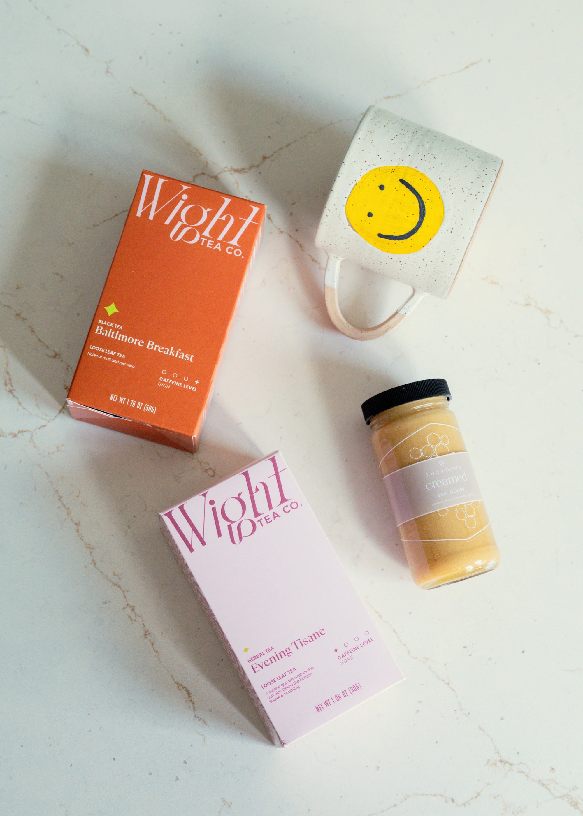 smiley face handmade mug, jar of creamed honey, and boxes of Wight Tea Co teas on a pale background.