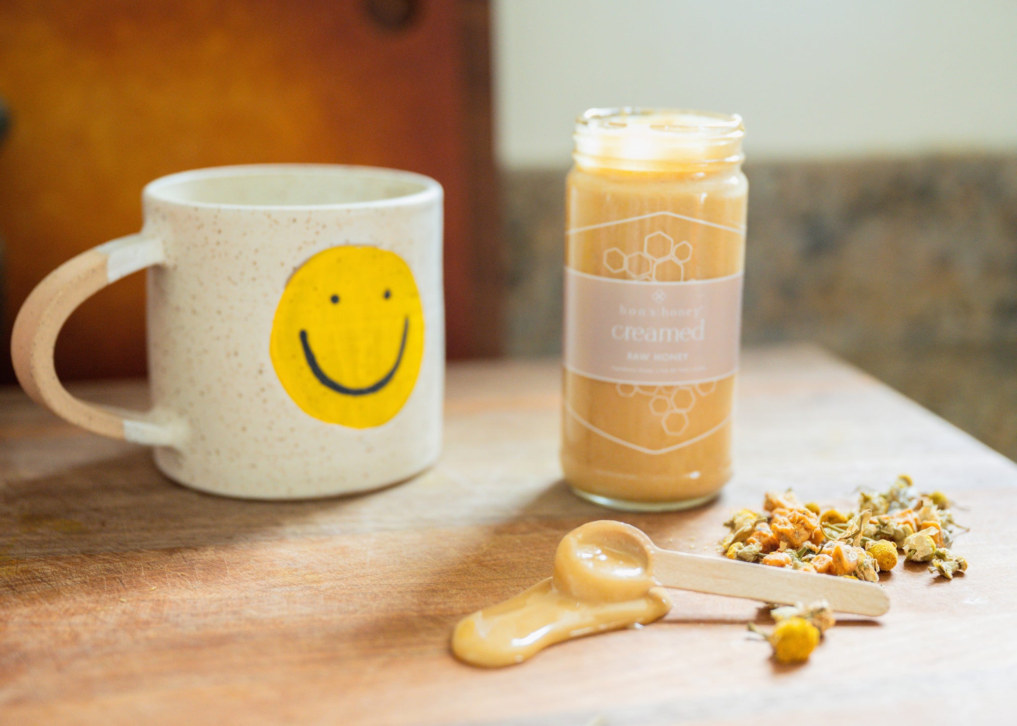 Mug with smiley face design, jar of creamed honey, and dried loose leaf tea on a wooden surface.
