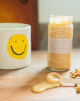 Mug with smiley face design, jar of creamed honey, and dried loose leaf tea on a wooden surface.