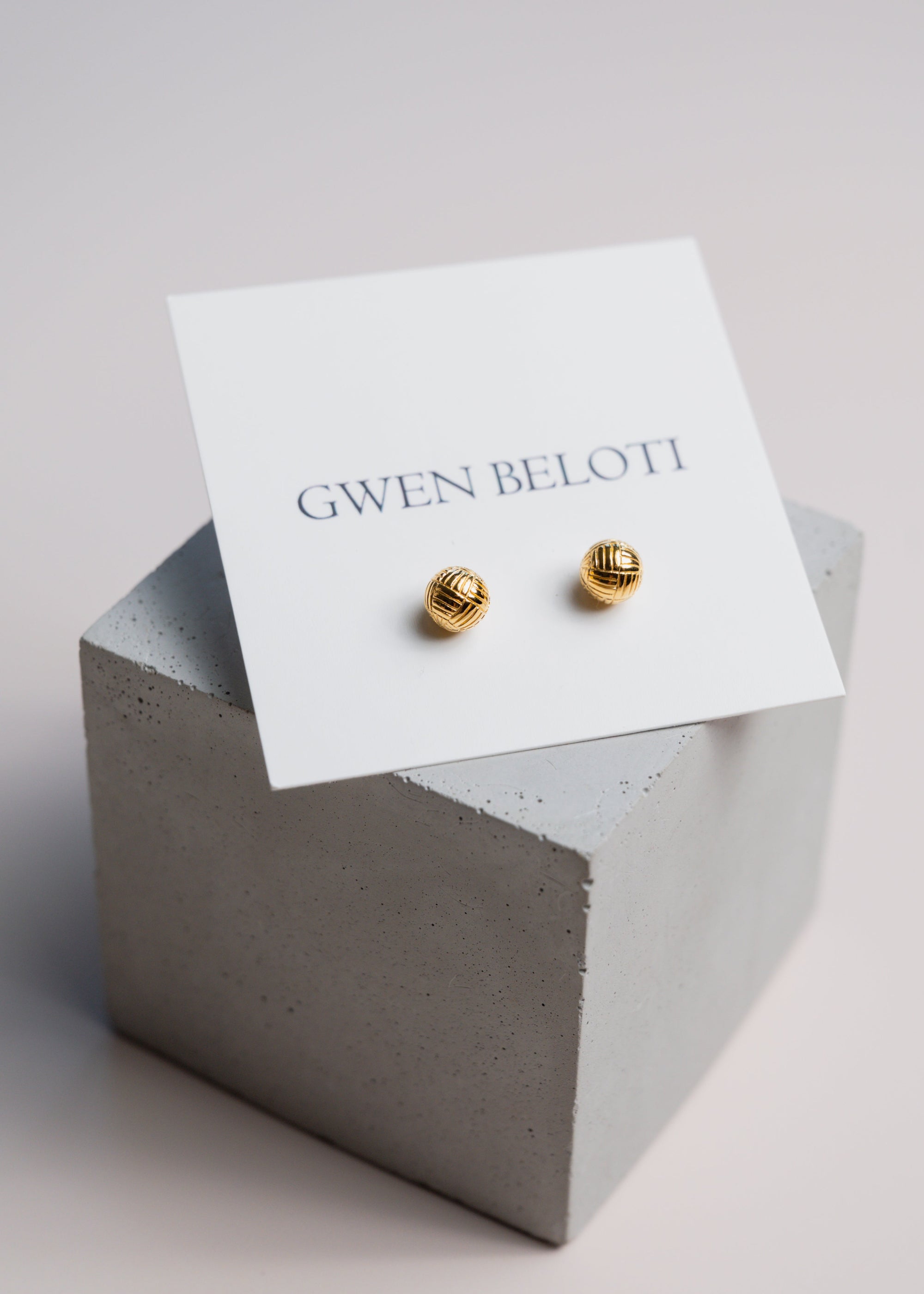Gold woven ball earrings by Gwen Beloti displayed on a concrete block.